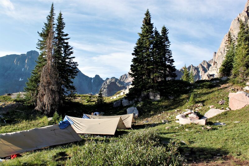 The image shows a campsite nestled in a mountainous landscape. Several tents are pitched on a grassy area, surrounded by evergreen trees. In the background, rugged mountain peaks rise against a blue sky with scattered clouds. The scene suggests a remote and peaceful outdoor setting.